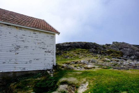 Téléchargez les photos : Olberg ; Olbergstranden ; Raege ; Norvège ; 20 mai 2023, Ancien bâtiment traditionnel avec un paysage rocheux en arrière-plan sous un ciel bleu brumeux - en image libre de droit