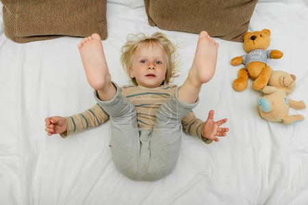 Foto de Cute little blond caucasian child kid toddler boy playing,having fun, lying on bed,lifts up the legs at home at sunny morning wakes up in his room. happy childhood moments. - Imagen libre de derechos