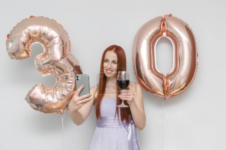 Happy young millennial attractive woman hold festive balloons number 30,glass wine,making selfie,accepts online greetings by phone,smiling celebrating her birthday at home holiday surprise party.