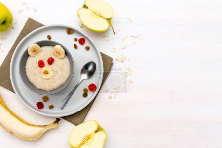 Funny cute kids childrens baby's healthy breakfast lunch oatmeal porridge in bowl look like bear face decorated with apple,banana,dried berry fruits. dessert food art on white wooden table.copy space.