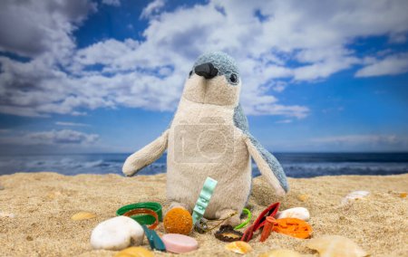 Photo for A penguin teddy surrounded by rubbish and trash on a beautiful beach - Royalty Free Image