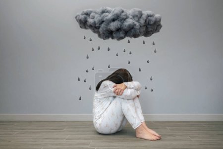 Mujer irreconocible con trastorno mental y pensamientos suicidas llorando sentada bajo una nube de tormenta oscura y gotas de lluvia cayendo sobre su cabeza en una habitación. Emociones negativas y malos sentimientos concepto.