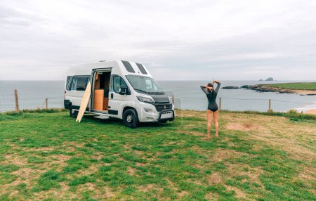 Unrecognizable young woman closing the zipper of her wetsuit for surfing next to her camper van