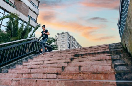 Foto de Feliz joven corredora entrenando escaleras abajo en pista urbana con palmeras al atardecer - Imagen libre de derechos