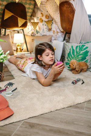 Little girl using walkie-talkie lying over rug surrounded by cushions and plants in teepee.Happy child playing with a walkie talkie in cozy shelter tent.Staycation and vacation camping at home concept