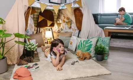 Little girl holding walkie-talkie lying over rug in cozy shelter tent while talk with boy sitting on couch in background. Two children playing with walkie talkies in teepee. Vacation camping at home.