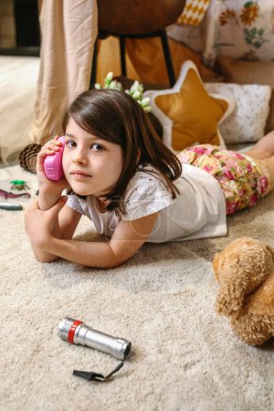 Portrait of little girl looking at camera while talking with toy phone lying over rug surrounded by cushions.Happy child playing with a plastic cell in cozy living room. Vacation at home concept.