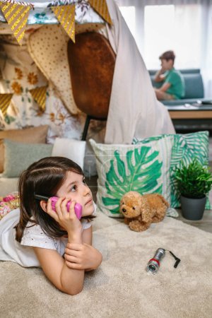 Pensive little girl holding walkie-talkie lying over rug in cozy tent while talk with boy sitting on couch. Two concentrated children playing with walkie talkies in teepee. Vacation camping at home.