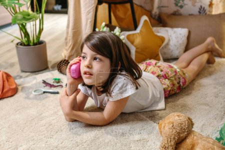 Portrait of little girl talking with toy phone lying over rug surrounded by cushions and plants in handmade teepee.Happy child playing with a plastic cell in cozy shelter tent.Vacation camping at home