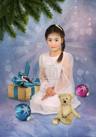 Téléchargez les photos : Petite fille en robe blanche assise sur ses genoux avec un ours en peluche entouré de cadeaux de Noël sur fond d'hiver - en image libre de droit