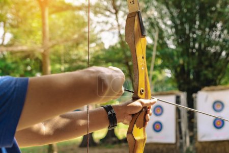 Photo for Hands of young man aims archery bow and arrow to colorful target in shooting range during training. Exercise and concentration with outdoor archery. Selective focus on hand. Sport, Recreation concept. - Royalty Free Image