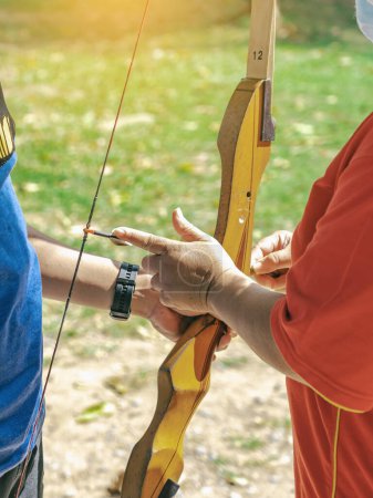 Photo for Female teacher teaches student to aim at goal. An archer teaching young man archery on field. Instructor teaching man to use bow and arrow on archery training. Archery course. Selective focus on hand. - Royalty Free Image