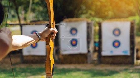 Photo for Hands of woman aims archery bow and arrow to colorful target in shooting range during training. Exercise and concentration with outdoor archery. Selective focus on hand. Sport, Recreation concept. - Royalty Free Image