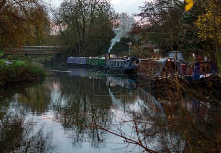 Photo for Sawbridgeworth, Hertfordshire England-November 27th 2022: A group of colourful canal boats moored at the Maltings mooring on the River Stort in Sawbridgeworth. A mallard also swims across the river. - Royalty Free Image