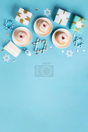 Hanukkah sweet doughnuts sufganiyot (traditional donuts) with fruit jelly jam, gift boxes, spinnig driedel and candles on blue paper background. Jewish holiday Hanukkah concept. Top view, copy space.