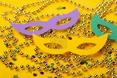 Photo for Mardi Gras gold color beads with Masquerade festival carnival masks and golden, green, purple confetti on yellow background. Party invitation, greeting card, venetian carnivale celebration concept. - Royalty Free Image