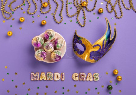 Téléchargez les photos : Mardi Gras King Cake cupcake or muffins, masquerade festival carnival mask, gold beads and golden, green confetti on purple background. Holiday party invitation, greeting card concept. - en image libre de droit