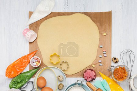 Foto de Sweet baking concept for Easter. Cooking background with shortbread dough, eggs, rolling pin, whisk for whipping, cookie cutters, sugar sprinkling, flour. Rustic white wooden background, top view copy space. - Imagen libre de derechos