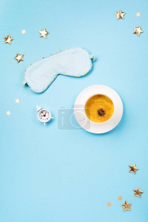 Foto de World Sleep day observed on March. Quality of sleep, good night, insomnia, relaxation concept. Sleeping mask, Turmeric golden milk latte and white alarm clock on blue background. Flat lay, top view - Imagen libre de derechos