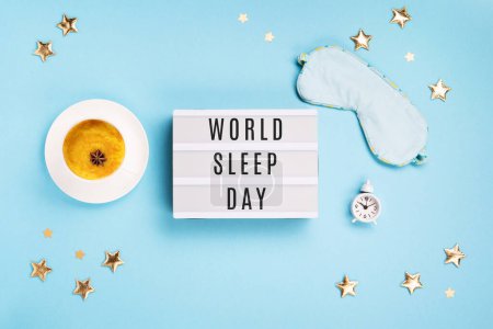 World Sleep day observed on March. Quality of sleep, good night, insomnia, relaxation concept. Sleeping mask, Turmeric golden milk latte and white alarm clock on blue background. Flat lay, top view