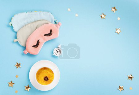 World Sleep day observed on March. Quality of sleep, good night, insomnia, relaxation concept. Sleeping mask, Turmeric golden milk latte and white alarm clock on blue background. Flat lay, top view