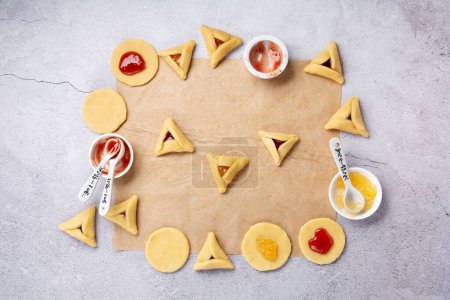 Téléchargez les photos : Making homemade Triangular pastry, Hamantaschen cookies for Purim. Purim celebration, jewish carnival holiday concept. Dough, jam, sugar, butter and rolling pin on gray stone table. Top view, flat lay, copy space. - en image libre de droit