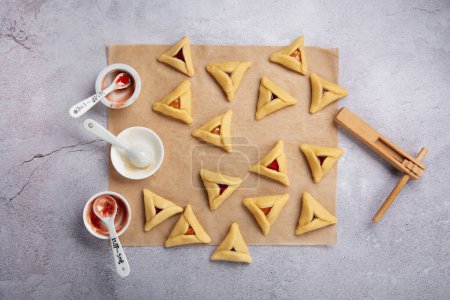 Téléchargez les photos : Making homemade Triangular pastry, Hamantaschen cookies for Purim. Purim celebration, jewish carnival holiday concept. Dough, jam, sugar, butter and rolling pin on gray stone table. Top view, flat lay, copy space. - en image libre de droit