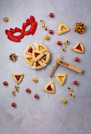 Foto de Purim celebration jewish carnival holiday concept. Tasty hamantaschen cookies, Triangular pastry, red carnival mask, noisemaker, sweet candies and party decor on gray stone table. Top view, flat lay, copy space. - Imagen libre de derechos