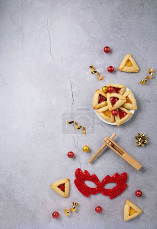 Purim celebration jewish carnival holiday concept. Tasty hamantaschen cookies, Triangular pastry, red carnival mask, noisemaker, sweet candies and party decor on gray stone table. Top view, flat lay, copy space.