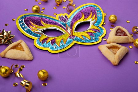 Photo for Purim celebration jewish carnival holiday concept. Tasty hamantaschen cookies, Triangular pastry, Festive carnival mask, noisemaker, sweet candies and party decor on purple Background, copy space. - Royalty Free Image