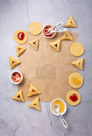 Foto de Making homemade Triangular pastry, Hamantaschen cookies for Purim. Purim celebration, jewish carnival holiday concept. Dough, jam, sugar, butter and rolling pin on gray stone table, Top view. - Imagen libre de derechos