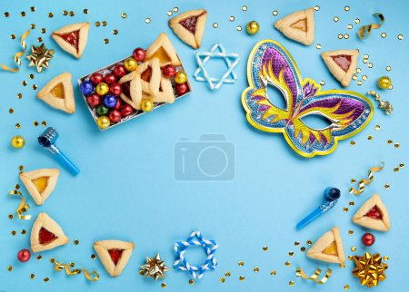 Foto de Purim celebration jewish carnival holiday concept. Tasty hamantaschen cookies, Carnival mask, noisemaker, sweet candies and party decor on blue background. Top view, flat lay, copy space. - Imagen libre de derechos