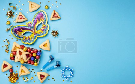 Photo for Purim celebration jewish carnival holiday concept. Tasty hamantaschen cookies, Carnival mask, noisemaker, sweet candies and party decor on blue background. Top view, flat lay, copy space. - Royalty Free Image