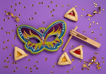 Foto de Purim celebration jewish carnival holiday concept. Tasty hamantaschen cookies, Triangular pastry, Festive carnival mask, noisemaker, sweet candies and party decor on purple Background. Top view, flat lay, copy space. - Imagen libre de derechos