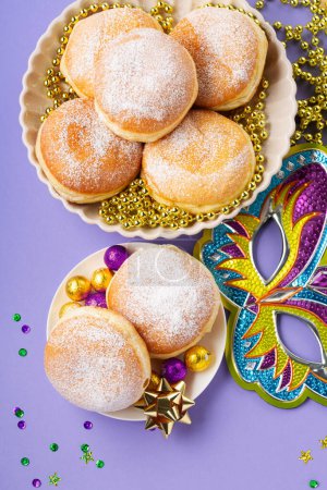 Téléchargez les photos : Mardi Gras King Cake doughnuts or donuts, masquerade festival carnival mask, gold beads and golden, green confetti on purple background. Holiday party invitation, greeting card concept. - en image libre de droit