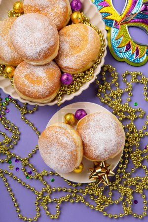 Foto de Mardi Gras King Cake doughnuts or donuts, masquerade festival carnival mask, gold beads and golden, green confetti on purple background. Holiday party invitation, greeting card concept. - Imagen libre de derechos