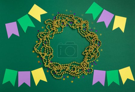 Photo for Mardi Gras masquerade festival card. Gold beads, colorful flags and golden, green, purple confetti on green background. Festive Holiday party invitation, greeting card concept. Top view, copy space. - Royalty Free Image