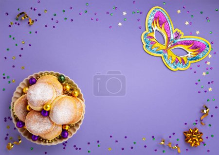 Téléchargez les photos : Mardi Gras King Cake doughnuts or donuts, masquerade festival carnival mask, gold beads and golden, green confetti on purple background. Holiday party invitation, greeting card concept. - en image libre de droit