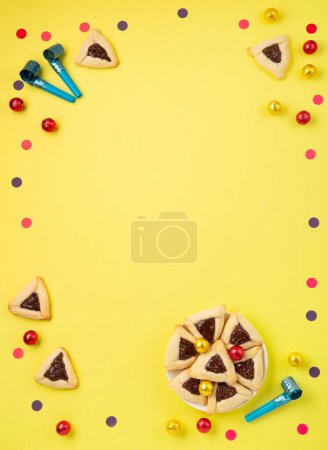 Téléchargez les photos : Homemade Purim hamantaschen cookies, Triangular pastry, Carnival mask, noisemaker, sweet candies and festive party decor on yellow background, Top view. Purim celebration jewish holiday concept. - en image libre de droit