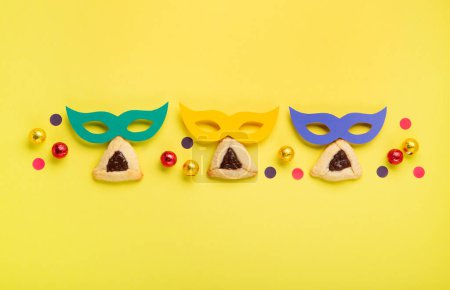 Foto de Homemade Purim hamantaschen cookies, Triangular pastry, Carnival masks, sweet chocolate candies and festive party decor on yellow background, Top view. Purim celebration jewish holiday concept. - Imagen libre de derechos