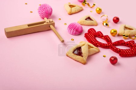 Téléchargez les photos : Purim celebration jewish carnival holiday concept. Tasty hamantaschen cookies, Triangular pastry, red carnival mask, noisemaker, sweet candies and party decor on pink background. Top view, flat lay, copy space. - en image libre de droit