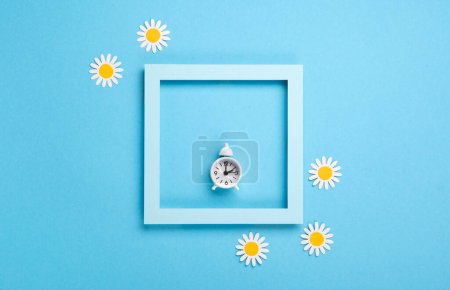 Téléchargez les photos : Réveil blanc et fleurs de marguerite sur fond bleu. Avance du printemps, Changement d'heure, Fin de l'heure d'été, Changement d'heure de la montre au printemps, Concept de retour d'été. - en image libre de droit