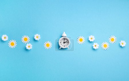 Foto de White alarm clock and Daisy Flowers on blue background. Spring forward, Time Change, Daylight Saving Time Ends, Changing the time on the watch to spring time, Summer back concept. - Imagen libre de derechos