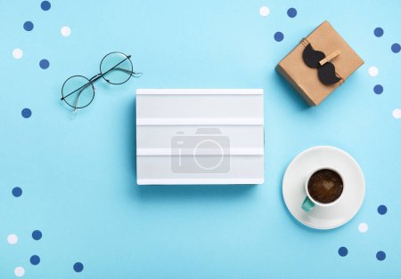 Photo for Fathers day holiday card concept with craft gift box, black moustache, cup of coffee, empty white lightbox and glasses on blue background, top view, copy space. - Royalty Free Image
