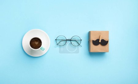 Téléchargez les photos : Fathers day holiday card concept with craft gift box, moustache noire, tasse de café et verres sur fond bleu, vue de dessus, espace de copie. - en image libre de droit