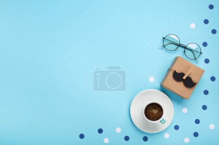 Foto de Concepto de tarjeta de vacaciones del día de los padres con caja de regalo artesanal, bigote negro, taza de café y vasos sobre fondo azul, vista superior, espacio para copiar. - Imagen libre de derechos