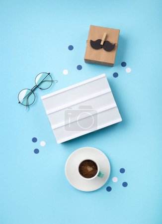 Téléchargez les photos : Fathers day holiday card concept with craft gift box, moustache noire, tasse de café, lightbox blanc vide et lunettes sur fond bleu, vue dessus, espace de copie. - en image libre de droit