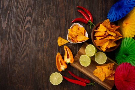 Photo for Cinco de Mayo (Fifth of May) celebration concept. Mexican background fiesta with Traditional Mexican nachos chips, red chili peppers, green lime on dark wooden background. Top view, copy space. - Royalty Free Image