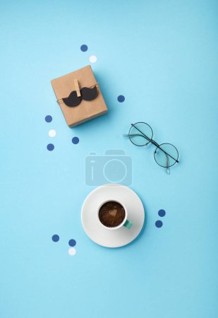 Photo for Fathers day holiday card concept with craft gift box, black moustache, cup of coffee and glasses on blue background, top view, copy space. - Royalty Free Image