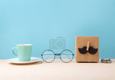 Photo for Fathers day holiday card concept with craft gift box, black moustache, cup of coffee, book and glasses on blue background, copy space. - Royalty Free Image